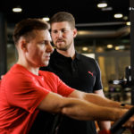 Ein Manhattan Fitnesstrainer unterstützt einen Mann im roten T-Shirt beim Krafttraining.